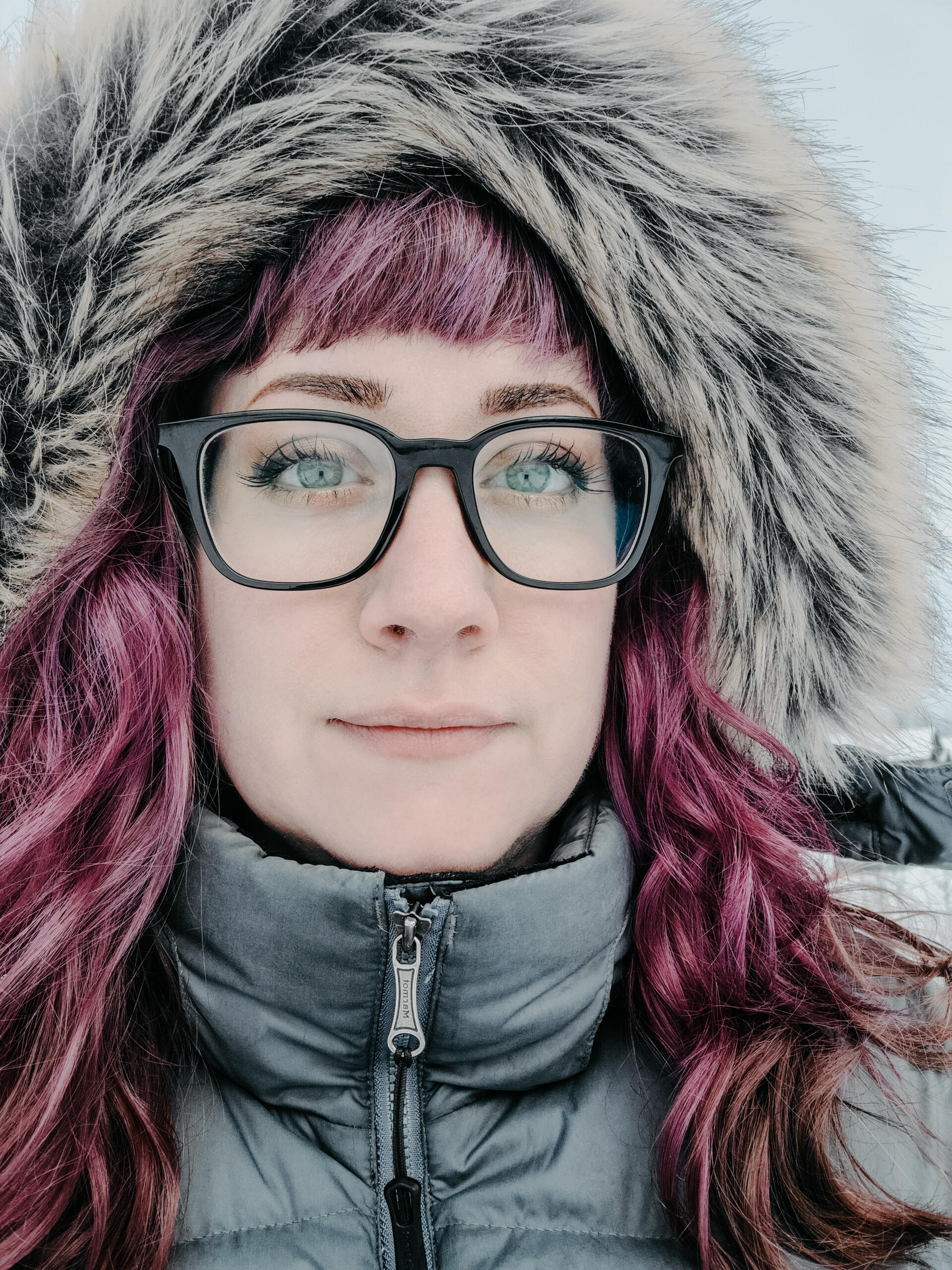 A photo of a woman with long brown hair and glasses looking away from the camera against a backdrop of corrugated metal.